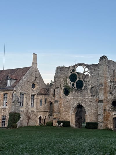 A partially ruined stone building stands adjacent to a building with a gabled, tiled roof and a prominent chimney. The ruined structure features large, circular openings and an arched entrance. Both buildings exhibit intricate stonework. Greenery is present in the form of a lawn and hedges near the buildings. The sky is visible in the upper left corner.