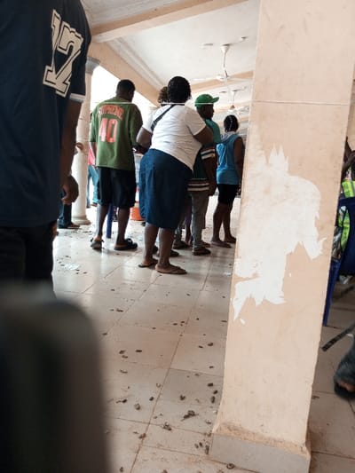 The image depicts an indoor or semi-outdoor public gathering space in Ajalli, Nigeria. The ground is a light-colored tiled floor, covered with numerous small, dark fragments resembling groundnut shells and several scattered white paper scraps. Two cream-colored square pillars are visible, one in the foreground right exhibiting significant peeling of paint or plaster. Overhead, at least three white ceiling fans are mounted.
In the immediate foreground on the left, the back of a person wearing a dark blue t-shirt with a large white graphic, appearing as "1Y" or similar, is present. In the midground, facing away from the camera, a man wears a green t-shirt with "SUPREME 40" printed on the back, black shorts, and dark sandals. To his right, a woman in a white t-shirt, a dark blue knee-length skirt, and light brown sandals carries a black shoulder bag. Adjacent to her, a man wears a green baseball cap, a light blue t-shirt with horizontal stripes, and light-colored trousers. Further to the right, a woman in a light blue sleeveless top and dark shorts is visible. Other partial figures are present in the deeper background. A white bucket or container is partially obscured behind the woman in the white t-shirt, and a blue plastic chair is visible on the far right edge of the frame. The individuals are standing, appearing to be queuing or gathering.