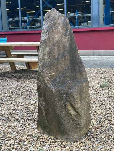 This media file depicts a large, rough-textured boulder lying on a bed of small, light-colored gravel. The boulder is dark gray with visible veins and variations in color and texture, suggesting it is a natural rock. Its shape is irregular, elongated, and somewhat flattened.
In the background, to the left of the boulder, is a building with a red wall. Behind the red wall, a large industrial shelving unit with blue and silver racks is visible, filled with what appear to be boxes or other stored items. Fluorescent lights are visible within the shelving unit, suggesting an indoor or covered industrial space. A pale grey concrete or stone surface is also visible between the red wall and the shelving unit.
To the right of the boulder, and extending towards the foreground, is more of the light-colored gravel. A small, green plant is growing out of the gravel near the top of the image. A wooden structure, possibly part of a bench or display, is visible behind and to the right of the boulder, constructed from light-colored wooden planks. A small, bright blue object, possibly a plastic crate or container, is partially visible near the bottom center.
The scene appears to be outdoors, likely in an industrial or landscaping area. The lighting suggests daytime, but the specific time of day and weather conditions are not discernible. The image is oriented vertically, with the boulder running horizontally across the frame. There is no text visible in the media file. The location context is Saran, France.
