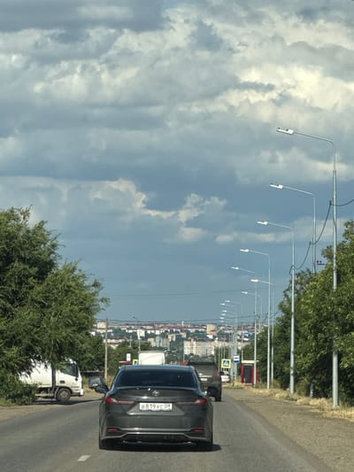 This image, taken roadside in Russia, depicts a cloudy day with a view of a cityscape in the distance. The foreground is dominated by a grey car with the license plate "B819XC 09" visible. To the left of the car, a white truck is parked. The road extends into the distance, lined with numerous streetlights that curve upwards and away from the viewer. Dense green trees line the right side of the road, bordering the asphalt. In the background, buildings of varying heights and colors form a town or city skyline. The sky is filled with dramatic, cumulus clouds, suggesting it might be midday or late afternoon. The overall scene conveys a sense of everyday travel along a suburban or semi-urban road.