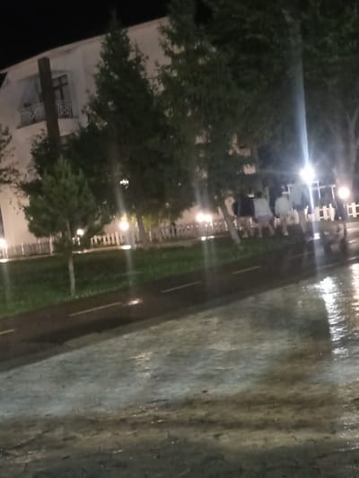 The image depicts a nighttime scene in the city of Sary-Oy, Kyrgyzstan. A multi-story building with balconies is visible in the background, partially obscured by trees. The foreground shows a wet, reflective surface, likely a road or pathway, indicating recent rain or damp conditions. Several individuals are walking or standing near the building, their figures blurred by the low light and possibly motion. Streetlights and lights from the building illuminate the scene. The overall atmosphere is dark and slightly hazy, consistent with a nocturnal urban environment.