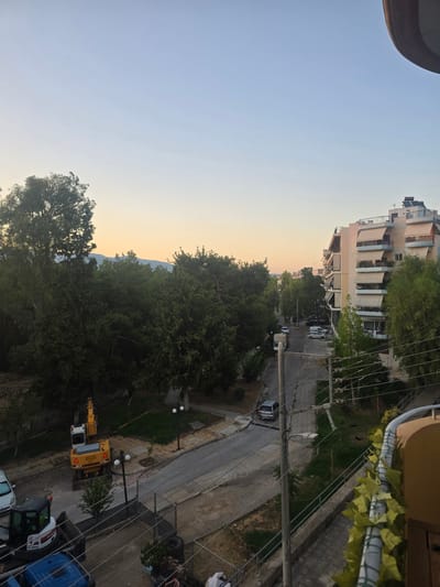 The image captures a street scene in Nea Filadelfeia, Greece, with a view from an elevated position, likely a balcony. The sky is clear and shows a gradient of light blue to a pale yellow near the horizon, indicating either early morning or late afternoon.
In the foreground, a balcony railing is visible, adorned with artificial green leaves. Below, a paved walkway leads down to a street. On the street, several vehicles are parked and in motion. A white excavator, identified by "LIEBHERR 902", is parked on the side of the road near some construction materials. To the left of the excavator, a smaller white construction vehicle, a Bobcat, is also present.
The street is lined with trees on both sides. To the right, a multi-story apartment building stands, characterized by its cream-colored facade and numerous balconies equipped with beige awnings. The building has a circular structure at the top with what appears to be solar water heaters. To the left, the landscape is dominated by dense green trees, with a large tree in the middle ground. In the distance, a range of hills or mountains is visible against the horizon.
Power lines and streetlights are present along the street, indicating urban infrastructure. The overall scene suggests a quiet residential area undergoing some minor construction work. There are no people clearly visible in the frame.
