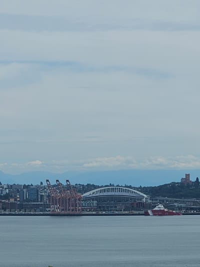 This is a photograph taken from across a body of water, likely Puget Sound, looking towards the Seattle skyline under a cloudy sky. In the foreground, the water is a calm, grey-blue color.
Dominating the middle ground are several large, coral-colored shipping cranes clustered together on the waterfront. Behind the cranes, a dense urban landscape unfolds with numerous buildings of varying heights and architectural styles. Among the buildings, a large, modern stadium with a distinctive white arched roof is visible, with its stadium seating clearly discernible. To the right of the stadium, a red and white ship, identified by the "U.S. COAST GUARD" lettering on its side, is present. Further in the background, a range of hazy blue mountains forms the horizon line. A solitary, reddish-brown brick tower structure with visible windows stands on a green, tree-covered hill on the far right side of the image.
In the upper portion of the image, the sky is a pale blue with scattered, soft white clouds. A very small, bright white dot, possibly an aircraft or a distant object, is visible in the sky above the stadium. The overall impression is a view of Seattle's port and waterfront area, with the stadium being a prominent landmark.