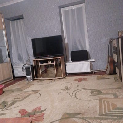 The image depicts an indoor scene in an apartment in Vasylkiv, Ukraine, likely a living room. A large flat-screen television sits on a wooden entertainment stand with glass doors. To the left of the television, a silver computer tower with cables is visible, placed on the floor. The room features a patterned carpet with floral motifs in shades of beige, green, and red. A radiator with white slats runs along the wall beneath a window draped with sheer white curtains. A black speaker is positioned on top of another radiator on the right side of the room. Further to the right, a guitar with a wooden body and neck leans against a piece of furniture. The walls are covered in textured wallpaper with a speckled, grey finish. The overall lighting suggests it is daytime, but the room appears dimly lit.