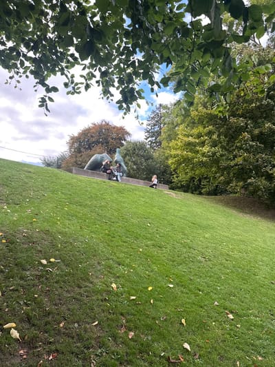 The scene is an outdoor park area within the Louisiana Museum of Modern Art in Humlebæk, Denmark. The foreground features a steeply sloping grassy knoll, covered in green grass with scattered fallen yellow and brown leaves. Overhanging green tree foliage partially obscures the sky at the top of the frame. Roughly midway up the knoll, a rectangular concrete plinth supports a large, dark, biomorphic abstract sculpture, identified as Henry Moore's 'Reclining Woman' (1956). Three individuals are situated on this plinth. Two individuals are seated to the left of the sculpture, facing each other; one wears a dark jacket, the other a dark jacket with light-colored trousers. A third individual, in light-colored clothing, is seated further to the right on the plinth, facing away from the other two. The background consists of dense trees with varying green and autumnal brown foliage, under a partly cloudy sky.