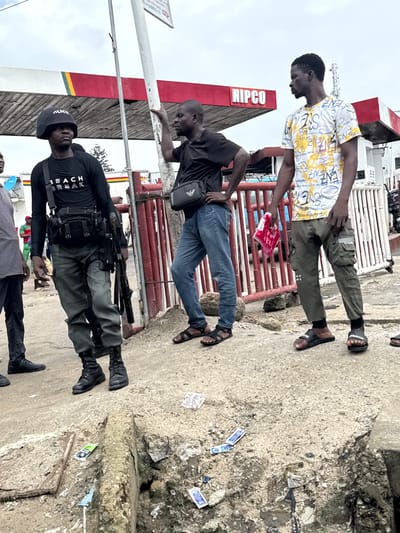 The image depicts a street scene in Lagos, Nigeria, with several people present. Two men are standing, one wearing a white t-shirt with yellow and black lettering and olive-green cargo pants, and the other in a black t-shirt and blue jeans. The man in the t-shirt and cargo pants appears to be handing a red item, possibly a packaged snack, to the man in the black t-shirt.
Another individual is visible further down, dressed in a black long-sleeved shirt with white lettering that appears to read "BEACH BREAK," a black tactical vest, and dark pants, with a rifle slung over his shoulder. He is wearing a black cap with "POLICE" written on it. The setting appears to be outdoors on a concrete surface, with some debris and discarded items on the ground, suggesting a public, possibly commercial, area. A red and white structure, possibly part of a building or signage, is visible in the background with the word "NIPCO" written on it. The sky is overcast, indicating a cloudy day. The overall atmosphere seems casual, with the interaction between the first two men appearing to be a simple exchange.