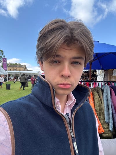 A young man is standing in front of a clothing stall at a market in Bramham, United Kingdom, a village located at latitude 53.871 and longitude -1.378. He is wearing a navy blue fleece vest with a brown leather trim, a pink shirt with a white checkered pattern, and his hair is parted on the side and brushed over his forehead. The man is looking directly at the camera with a serious expression on his face. The clothing stall behind him is displaying a variety of colorful jackets, sweaters, and other clothing items on hangers. There is also a blue and white striped umbrella in the background. The scene takes place outdoors, under a blue sky with white clouds. There are other people in the background walking past and some stalls, but they are out of focus. The scene is captured during the day in bright natural light.