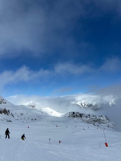 The image displays a detailed view of a snow-covered ski resort area in Bad Hofgastein, Austria, under a dynamic sky. The upper two-thirds of the frame is dominated by a deep blue sky, featuring scattered white clouds and faintly visible stars.
The foreground and midground present a vast, white ski slope covered in groomed snow with visible ski tracks. Several individuals are present: on the far left, a skier in a dark suit stands; in the center-left, another skier in a yellow jacket and dark pants descends the slope; and further right, a third skier in an orange jacket is visible. Numerous orange pole markers with square tops delineate areas of the slope, accompanied by a blue circular marker with a white symbol on the snow.
In the midground, various snow-covered mountain peaks and ridges are visible. A multi-story building with a brown roof, likely a mountain restaurant or station, is situated on a distant peak, left of center. To the right, a rocky outcrop with scattered pine trees and additional structures can be observed. The background features larger snow-capped mountains partially obscured by thick white clouds that extend across the horizon, blending with the sky. The scene depicts active winter sports conditions with multiple skiers on the designated slopes.
