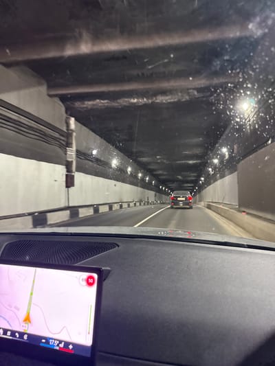 The image shows the view from inside a vehicle, looking into a tunnel. The tunnel has a gray and white color scheme, with reflective lights lining the walls. The vehicle's navigation system is visible, displaying a map with an arrow indicating direction. The map details appear to show "Sevan-Dilijan Tunnel" and "M4" and "Hrazdan River" suggesting the vicinity of Semyonovka, Armenia. The speed indicated on the navigation system is 69 km/h. The windshield appears to have raindrops on it. A gray interior panel of the vehicle is visible to the right of the view.