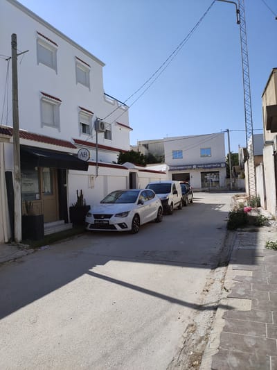 The image depicts a street scene in Cebalat, Tunisia, under a clear blue sky. In the foreground, a white sedan is parked on the asphalt road, with its front license plate displaying "213 8604". To the left of the car is a building with a white facade, featuring a black awning over an entrance and a circular sign with the letters "AB" in black. Further down the street, another white building has signage that reads "VENTE EAU MINERALE EN GROS" and "بيع المياه المعدنية بالجملة", indicating a wholesale mineral water business. Multiple vehicles, including vans and cars, are parked or driving along the street. The environment includes paved sidewalks, some vegetation, and a tall utility pole with wires. The buildings are of modern architecture with white walls and tiled roofs. The overall impression is of a quiet commercial or residential street.