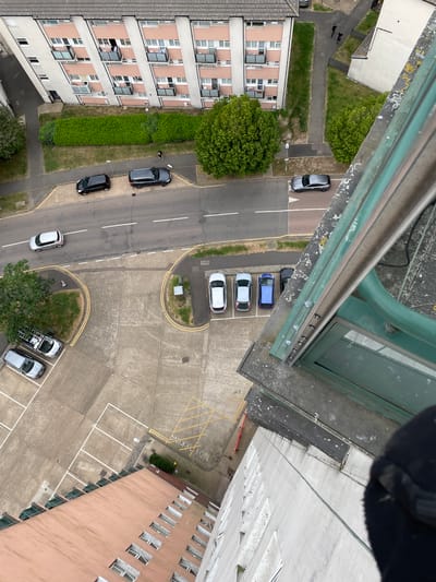 The image is a high-angle view, likely taken from a tall building, looking down on a street and adjacent parking areas in Hertsmere, United Kingdom. A multi-story apartment building with light-colored walls is on the left side of the image, and another building, possibly a commercial structure, is on the right, featuring windows on each floor.
A two-lane road runs diagonally through the center of the frame, with several cars parked along the sides. Yellow lines demarcate the parking spaces in the parking lots. A patch of green vegetation and trees is seen in the bottom center.
Two people are walking alongside the building on the left. One is holding a dog. There's a patch of lawn between them and the building. The immediate foreground shows part of a window structure and a blurred, dark-colored object that might be a gloved hand or part of clothing, suggesting the point of view is from a high window or balcony.