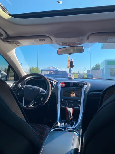 The image shows the interior of a Ford Fusion car parked outdoors under a clear blue sky. The car has a panoramic sunroof, which is partially open. The dashboard features a steering wheel with a Ford logo, a center console with a touchscreen display, and a gear shifter. The interior is upholstered in black with red stitching on the seats and gear shifter. There are also air fresheners hanging from the rearview mirror. The exterior view through the windshield shows a road, other vehicles, a building with a green roof, and trees. The location is identified as Herasymivka, Ukraine. The image appears to be taken on a sunny day.