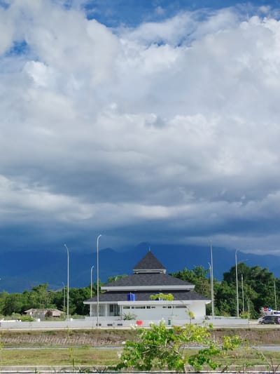 A modern white building with a dark, pyramidal roof, likely a mosque, is centered in the mid-ground of the image. The building features white pillars and a dark blue water tank on its roof. In front of it, a road runs horizontally, with a few vehicles, including a white van and a dark colored car, visible. A white wall borders the road and the building. To the left of the main building, there is a smaller, older-looking structure with a thatched roof and visible construction elements. Lush green trees form a dense background behind the buildings, and a range of dark blue mountains are visible in the far distance under a dramatic sky. The sky is filled with a mix of bright white and dark grey clouds, suggesting changing weather. The foreground shows a grassy area with some plants and white flowers. The overall scene depicts a semi-urban or semi-rural landscape in Keureuweungkrueng, Indonesia, under a dynamic sky.