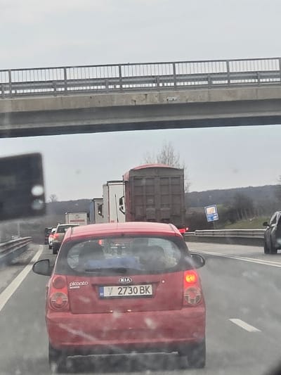 The image displays a multi-lane highway with stationary or slow-moving traffic under an overcast sky, observed from within a vehicle. Directly in front is the rear of a red 5-door Kia Picanto with Bulgarian license plate Y 2730 BK, its left rear taillight illuminated. Behind the Kia, a dark brown dump truck is visible, followed by a queue of other vehicles including white semi-trailer trucks and passenger cars. A concrete overpass with a silver metal railing spans horizontally across the upper third of the frame. A blue and white informational sign is present on the right shoulder of the road. The roadside vegetation consists of sparse, bare trees and shrubs, with distant hills visible under the grey sky. This scene is located on a major highway in the general vicinity of Plodovitovo, Bulgaria.