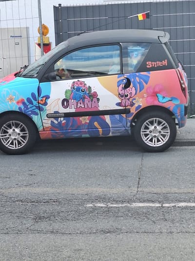 The image shows the side of a small car parked on a street in Bochum, Germany. The car is decorated with a colorful wrap featuring characters from Disney's Lilo & Stitch, including Stitch wearing a flower lei and Angel. The word "OHANA" is written in large pink letters across the side of the car, next to Stitch. The car also has the word "Stitch" written in pink lettering on the rear quarter panel. The car is parked on an asphalt road with visible cracks. In the background, a gray metal fence and a white building with a Panasonic sign are visible. A small German flag is attached to a wire above the car. The lighting suggests it is daytime.