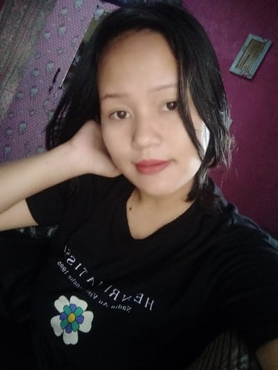A young woman poses for a selfie. She has black hair and brown eyes, and is wearing a black t-shirt with a white floral design and writing on the front. Her head is tilted slightly, and one hand is resting on her face. The background includes a patterned wall and a purple wall with a small rectangular object hanging on it. She is wearing lipstick.
