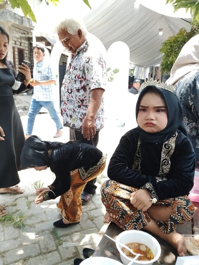 This image captures an outdoor gathering in Juwana, Indonesia, likely a celebratory event, under a white fabric canopy.
In the foreground, a young child, appearing to be a girl, sits cross-legged and barefoot on a wooden surface to the right. She wears traditional Javanese attire consisting of a black velvet long-sleeved top with gold embroidery, a black hijab, and a brown and gold batik sarong. A silver crown or tiara rests on her head, and she has visible makeup, including pink eyeshadow. Her expression is a distinct pout. Before her is a white plastic bowl containing a soup with meatballs, accompanied by a white plastic spoon. To her left, another child, similarly dressed in a black top, black hijab, and batik sarong, is bent over, facing away from the camera, attending to something on the paved ground.
In the middle ground, an older man with white hair stands upright, looking downwards and to his left. He wears a short-sleeved white batik shirt patterned with colorful birds, elephants, and foliage, paired with dark trousers. To his left, a young woman in a black long-sleeved dress holds a black smartphone horizontally, seemingly capturing an image or video. Further in the background, another man in a light blue patterned shirt and blue jeans walks away.
The environment features a light-colored paved ground with sparse patches of grass. Overhead, a white fabric canopy is visible, characteristic of outdoor event shelters. Two decorative white arched structures, possibly an entrance or photo backdrop, stand in the mid-ground. A building with exposed brick walls is visible in the background left, and green tree foliage is present at the top left and right edges of the frame. The event appears to be well-lit by natural daylight.