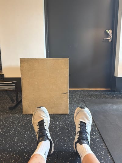 The image shows a first-person view of a person's feet wearing white athletic socks and white and black running shoes. The person is sitting on a gym floor, which is a speckled black surface. In front of the person is a wooden plyometric box. To the left of the box, part of a rowing machine is visible. Behind the box is a dark grey door with a silver handle and lock mechanism. The background also includes a plain light-colored wall. The scene is indoors, likely in a gym. The location in Östersund, Sweden, is not specified in the image.