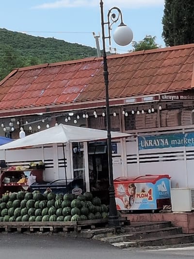 The image shows a street scene in Şəki, Azerbaijan, under a clear sky. The foreground features a lamp post with a white spherical lamp. To the right of the lamp post, a market stall is visible. This stall has a tiled red roof and displays various items for sale, including a large crate filled with watermelons. Behind the stall, a building with a white facade and blue accents can be seen. The building has signage that appears to be in Ukrainian, with text that reads "ukrayna market" and "UKRAYNA market". There is also a box with a depiction of children and the word "PLOM 1" and "AMELASS" written on it. The overall impression is of a lively marketplace on a sunny day.