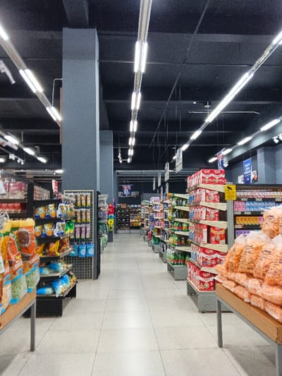 Inside a supermarket, rows of shelves stretch down an aisle, stocked with various goods. On the left, chips and snacks are displayed. The right features stacks of SGM products and bags of snacks. Overhead, fluorescent lights illuminate the scene. Gray pillars support the ceiling. Tiled flooring runs the length of the aisle.