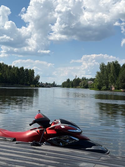 The image depicts a red and black jet ski parked on a wooden dock. The jet ski is positioned in the foreground, angled towards the right side of the frame. The water is calm, with gentle ripples reflecting the sky. In the background, a tree-lined shore stretches into the distance, with houses and buildings visible nestled amongst the trees. The sky is partly cloudy, with fluffy white clouds against a bright blue backdrop. The overall scene suggests a peaceful summer day at a lake or river, likely in a suburban or rural area. The location is the Klyazma River in Shchelkovo, Russia, a popular spot for water recreation.