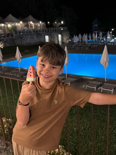 A young boy smiles while holding a watermelon-shaped popsicle in front of a brightly lit swimming pool at night. The boy is wearing a brown t-shirt and light-colored pants. In the background, there are lounge chairs, umbrellas, and buildings with lights. The setting appears to be a resort or hotel in Cavriglia, Italy.