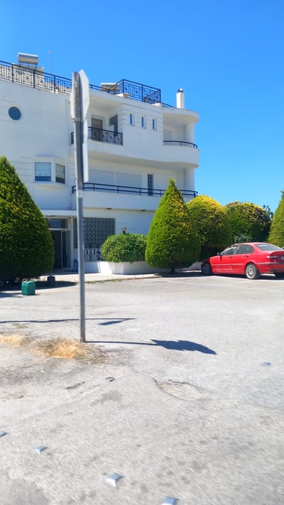 In the city of Lambi, Greece, an exterior scene is captured on a clear, sunny day with a bright blue sky. A white, multi-story building with balconies and solar water heaters on the rooftop dominates the background. In front of the building, a red sedan is parked. Several meticulously manicured evergreen trees and bushes are planted in front of the building. The foreground consists of a paved area, likely a street or parking lot, with some scattered debris and reflective markers on the surface. A traffic signpost is partially visible in the middle ground. A green plastic container is situated on the ground near the left side of the frame.