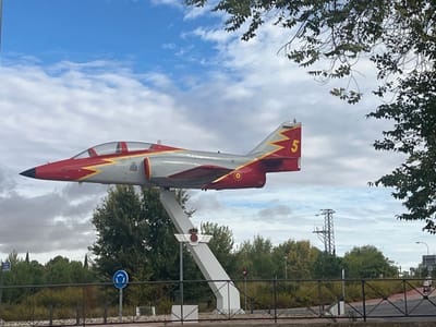The image displays a static outdoor monument featuring a Spanish Air Force CASA C-101 Aviojet aircraft mounted horizontally on an angled white pedestal. The aircraft is painted primarily in grey and red, with a red nose cone, red canopy frame, red underside of the fuselage, and a red tail section. A yellow zig-zag stripe runs along the fuselage, and a yellow number "5" is prominently displayed on the tail fin. A Spanish Air Force roundel is visible on the lower rear fuselage. The white pedestal, likely made of concrete or painted metal, supports the aircraft and features a crest surmounted by a crown.
The immediate foreground includes a black metal fence. Behind the fence, to the left of the pedestal, a blue circular road sign with a white arrow indicating a mandatory roundabout is visible. The middle ground is dominated by a variety of green trees and shrubs, some showing autumnal yellowing. In the far background, a large metal lattice electricity pylon stands against a sky characterized by scattered white and grey clouds on a blue expanse. No individuals, actions, or direct interactions are depicted in the scene.
This monument is situated in Alcalá de Henares, Spain, likely at a public intersection or entry point.