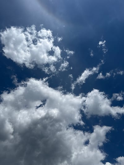 The image shows a bright blue sky with scattered white and grey clouds. In the upper left quadrant of the sky, a faint arc of a rainbow is visible. In the lower half of the image, the clouds are denser and appear to be darker, suggesting they may be cumulus clouds. There are a few small dark specks in the sky that could be birds or insects. The overall scene is one of a partly cloudy day. The image is a photograph taken outdoors, looking upwards at the sky. There are no visible structures or human figures in the frame. The photograph does not provide any information about the location in the city of Bucha, Ukraine.