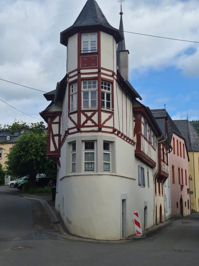 The image displays a daytime street scene in Traben-Trarbach, Germany, centered on a multi-story corner building. This prominent structure features a white facade with dark reddish-brown half-timbering (Fachwerk) and a distinctive rounded corner capped by a conical slate roof turret. A spire with a small weather vane or cross adorns the apex of the turret. On the upper section of the turret, a plaque reads "W. M." above "19 01", indicating a date. The building has numerous rectangular windows with white frames, some of which show white lace curtains. A set of closed blue shutters is visible on a lower window on the right side of the building. Rain gutters and downspouts are affixed to the facade. The lower part of the corner section shows minor peeling of paint.

To the immediate right of the main building, a light peach-colored building with dark reddish-brown window surrounds extends down the street. Further beyond, a pale yellow building is partially visible, featuring an exterior wall lamp. Both adjacent buildings appear to have dark slate roofs.

To the left of the main building, partially obscured by a large, leafy green tree, are other residential or commercial structures, including a yellow building with what appears to be solar panels on its roof. Two vehicles are parked on the left side of the street: a white hatchback and a dark SUV. A dark trash bin is positioned behind the corner of the main building on the left.

The asphalt road surface curves to the left and descends to the right. A raised sidewalk separates the buildings from the road. A circular metal manhole cover is embedded in the road surface, near which blue chalk markings are visible. Leaning against the front of the main building, on the street side, is a red and white striped temporary warning barrier or sign. Overhead, multiple dark electrical or communication wires traverse the upper portion of the frame against a sky that exhibits patches of blue interspersed with white and gray clouds. No people are visible in the scene.