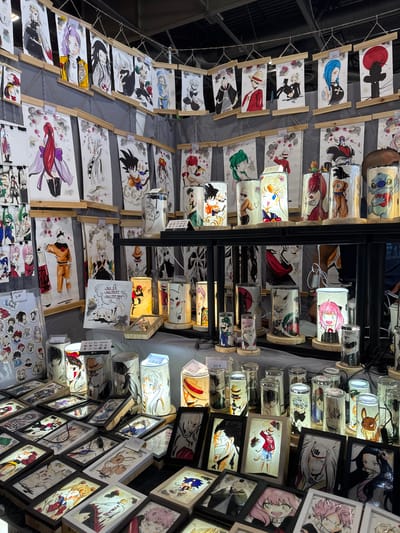 The image shows a stall at the Paris-Villepinte Exhibition Centre, likely during an anime or manga convention. The stall displays a variety of artworks, primarily anime-inspired prints and illuminated cylindrical lamps featuring characters from popular series like Dragon Ball Z, Sailor Moon, and My Hero Academia. Many of the prints are framed or mounted on wooden dowels and hung in rows. The lamps are arranged on tiered shelves, each lit from within to highlight the artwork. In the foreground, there are framed art pieces and a collection of smaller circular stickers or magnets. The overall setting is a retail environment with artificial lighting and a dark background.