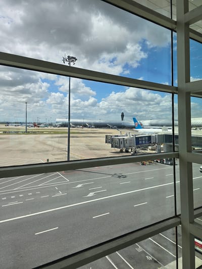 The image shows an airport tarmac viewed through a large glass window. In the foreground, a gray asphalt road is marked with white lines, indicating lanes and parking spaces. Beyond the road, the airport tarmac stretches out, with various airplanes visible, including a large white passenger jet with a blue tail logo, connected to a jet bridge. Several other aircraft are parked in the distance. The background features a modern airport terminal building with a distinctive curved, patterned roof. A tall control tower is visible to the left of the terminal. The sky is partly cloudy with a mix of blue sky and white cumulus clouds. The scene appears to be a sunny day at an international airport. The location is identified as Bao'an District, China.