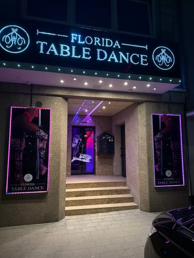 The image shows the exterior of a business called "Florida Table Dance" located in Duisburg, Germany. The entrance is illuminated by neon lights and features a sign with the business name. Two large posters advertising "Florida Table Dance" with images of dancers are displayed on either side of the entrance. The entrance itself is a doorway with stairs leading up to it. Inside, a dimly lit interior with purple and pink lighting is visible, along with what appears to be a vending machine. A black car is parked in front of the building. The street is paved with gray tiles, and the building facade is made of stone.