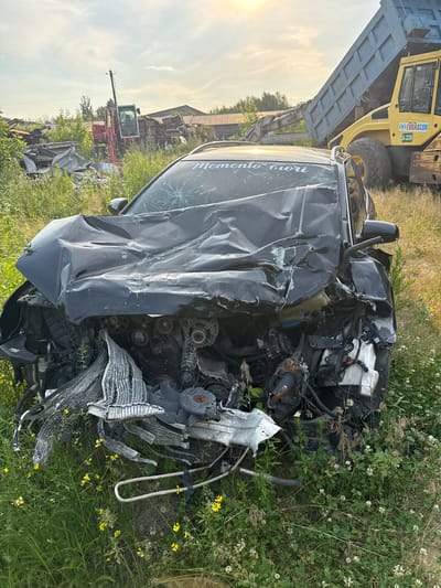 The image shows a severely damaged black car on its side, surrounded by overgrown green grass and vegetation with scattered white and yellow wildflowers. The car's front end is completely destroyed, revealing its engine and various mechanical components. The hood is heavily crumpled, and the windshield is cracked. The words "memento mori" are visible in white lettering on the car's body.

In the background, a large yellow construction vehicle, possibly a dump truck, is partially visible. To the left of the car, a red excavator is also present, with its arm extended. The scene appears to be an outdoor junkyard or a rural area, suggested by the presence of construction equipment and piles of debris. The lighting indicates it is daytime, with soft sunlight suggesting either early morning or late afternoon. The overall mood of the image is one of destruction and neglect, with the natural elements beginning to reclaim the man-made wreckage.