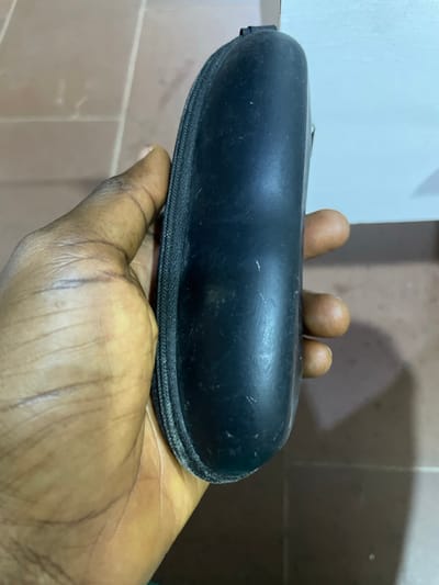 A dark-skinned human left hand holds an elongated, oval-shaped black zippered glasses case. The case exhibits a matte finish with visible surface dust and minor scuffs. The thumb is positioned on the left side of the case, while the fingers grasp the right side. The background features light brown square floor tiles with darker grout lines on the lower left and a lighter, possibly white or off-white, vertical surface on the upper right. The scene is illuminated by ambient light. This image was captured in Nibo, Nigeria.