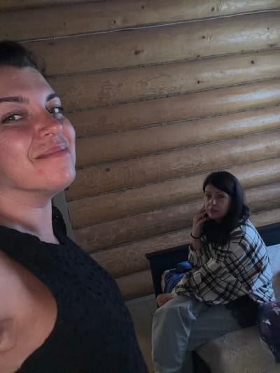 The image shows two individuals in a room with log walls. In the foreground, a woman with dark hair pulled back is looking towards the camera with a slight smile. She is wearing a black, textured top. In the background, another woman with dark hair is seated and holding a phone to her ear, looking off to the side. She is wearing a plaid shirt and light-colored pants. The room appears to be dimly lit, and the walls are constructed from horizontal logs. The location is identified as Шапки, Russia.