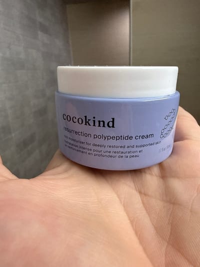 A Cocokind Resurrection Polypeptide Cream jar is held in a person's left palm. The jar is light purple with a white lid and black text that reads "cocokind resurrection polypeptide cream rich moisturizer for deeply restored and supported skin hydration intense pour une restauration et un renforcement en profondeur de la peau 1.7 fl oz / 50 ml". The background appears to be a tiled wall and a neutral-colored wall. This image was taken in Burbank, United States.