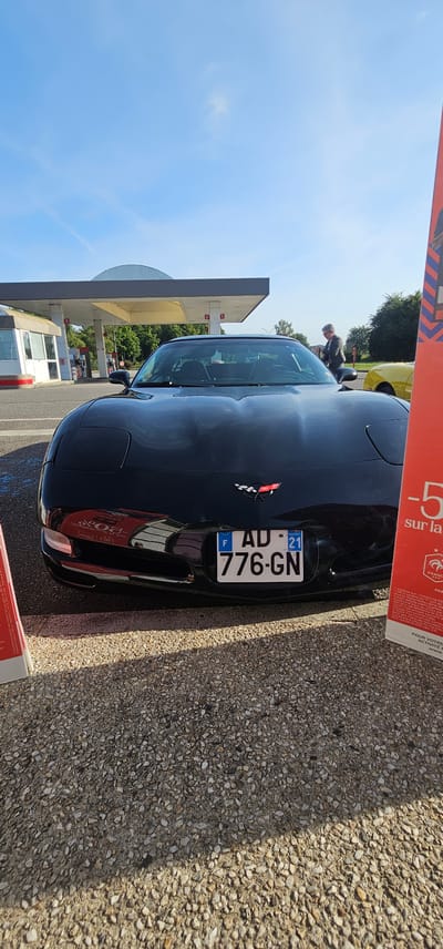 A black, fifth-generation (C5) Chevrolet Corvette is positioned centrally, viewed from the front, parked on a light-colored gravel surface at a gas station in Bain-de-Bretagne, France. The vehicle's front bumper displays a French license plate, "AD-776-GN," with the departmental identifier "21" (Côte-d'Or) on the right-hand blue band. The checkered flag Corvette emblem is visible on the center of the front fascia. The glossy black hood reflects a partial red advertising stand visible on the right side of the frame.

In the background, a gas station facility is visible, featuring a grey and white canopy covering fuel pumps. Pump numbers "1," "2," and "5" are partially discernible on the canopy's support columns. To the far left, a white building, possibly a kiosk or service shop, is present. A yellow car is parked further back on the right side of the gas station, and an adult male with short grey hair, wearing a patterned jacket, is standing near it, facing towards the pumps. Green foliage and trees form a dense background behind the gas station structure. The sky above is clear and blue, exhibiting faint contrails. Strong sunlight illuminates the scene, casting distinct shadows, notably from the foreground advertising stand onto the gravel.