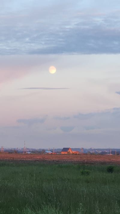The image depicts a landscape at dusk or dawn in the vicinity of Бельково, Раменский район, Russia. The sky occupies the majority of the frame, transitioning from a pale blue with cumulus clouds on the left to a muted pink and light orange towards the center. A bright, round object, likely the moon or sun, is visible through the clouds on the left. A thin, dark vertical cloud formation is positioned near the center.
The lower right portion of the image shows a field of green grass. Adjacent to the grass, a reddish-brown strip likely indicates a dirt path or road. Along this strip are sparsely distributed structures resembling houses or sheds, illuminated with an orange hue, suggesting either early morning light or artificial lighting. Utility poles are also visible along the road, spaced intermittently.