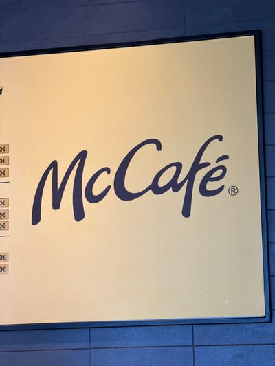 The image shows a sign for McCafé in Húmera-somosaguas-prado Del Rey, Spain. The sign is light yellow with dark blue text and a dark blue border. The McCafé logo is prominently displayed in the center of the sign. To the left of the logo, there are vertical lines of text with prices, indicating menu items and their costs. The sign is mounted on a dark blue tiled wall. There are no people visible in the image.