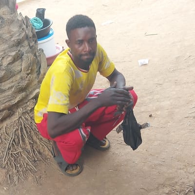 The image captures a full-length shot of a man sitting on a dirt ground, looking directly towards the viewer. The image appears to be rotated 90 degrees clockwise from its original orientation.
The man has dark skin, short dark hair, and a short beard. He is wearing a bright yellow short-sleeved t-shirt with a red and dark collar, which has some lighter discolored or patterned areas on the front. He is also wearing red track pants or similar casual trousers that feature white stripes running down the sides of the legs. On his feet, he wears dark-colored open-toed sandals or slides with lighter brown soles. His posture is relaxed, with his knees bent and drawn up towards his chest, and his hands clasped or resting on his lower legs. He has a neutral expression on his face.
The setting appears to be outdoors, with the ground composed of light brown, dry dirt or sand. Various small pieces of debris are scattered across the ground, including a small piece of white paper or plastic near the top left of the frame. To the left and slightly behind the man, there is a prominent section of a large, gnarled tree trunk or root system, light brown in color with a very rough, textured surface, and numerous thin, exposed fibrous roots extending outwards into the dirt. Next to this tree root, a white plastic bucket with a blue rim is visible, containing a black plastic bag and some light blue fabric or material. Behind the man's right leg, a black object resembling a folded umbrella or another black plastic bag rests on the ground.
The lighting in the image is bright and even, suggesting it was taken during daylight hours, possibly under an overcast sky or in a shaded area, as there are no harsh shadows. There is no discernible text visible within the image. The man appears to be resting or observing his surroundings.