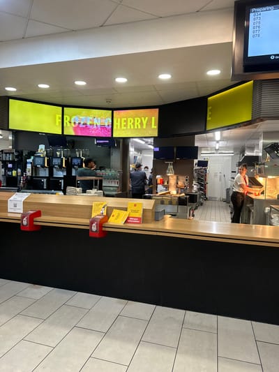 The image provides a detailed view inside a McDonald's fast-food restaurant, likely in Test Valley, United Kingdom, focusing on the service counter and kitchen area. The scene depicts a busy operational environment with several employees.
In the foreground, a long, light wood-toned service counter features black paneling along its base. Placed on the counter are red plastic holders, along with booklets titled "ALLERGEN information" and branded "McDonald's UK." Behind the counter, various machines, likely for beverages and dispensing, are visible, along with multiple smaller screens and payment terminals.
Several employees are actively working behind the counter in the kitchen. One individual, dressed in a white shirt and dark trousers, is preparing food at a station equipped with fryers and other stainless steel kitchen appliances. Other staff members are seen further back, engaged in food preparation tasks. The kitchen area appears clean and well-lit.
Digital display screens are prominent throughout the space. One large screen advertises "FROZEN CHERRY L." To the left, another screen displays "Preparing..." followed by a queue of order numbers: 034, 073, 075, 076, 077, and 078, indicating active orders awaiting completion. The ceiling is fitted with numerous recessed circular lights, providing bright illumination. The floor is tiled with light grey rectangular tiles. The overall setting suggests a typical fast-food operation during regular business hours.