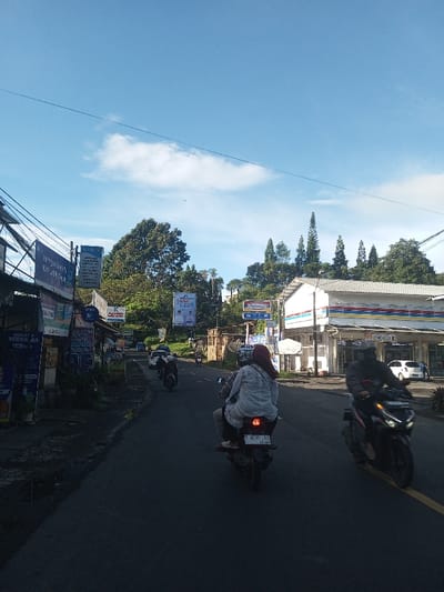 The image captures a street scene identified as being in the city of Khouathineung, Laos. The environment is sunny, with a clear blue sky and scattered white clouds. The asphalt road is populated with several motorcycles. In the foreground, a dark motorcycle with two occupants, a driver in dark clothing and a passenger wearing a light-colored top and a reddish head covering, proceeds forward. Its license plate, reading 'F 2C35 33', is visible. To the right, another dark motorcycle with a single rider in a dark jacket is in motion. The right side of the road features a white building with blue and yellow striped accents, characteristic of an Indomaret convenience store, complete with its visible signage. On the left side, a series of roadside businesses are present, displaying various signs, some with text in Indonesian, including one for an 'Apotek' (pharmacy) and campaign banners showing two men. Lush green trees and vegetation cover hillsides in the background, with some tall, slender trees prominent. Overhead utility lines are visible on the left.