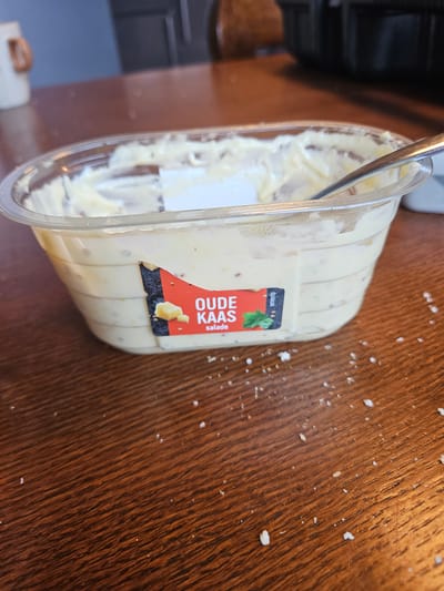 An overhead, slightly angled medium shot captures a clear plastic tub of "Oude Kaas Salade" (Old Cheese Salad) on a brown wooden table. The tub is partially full, with a silver spoon inserted into the creamy yellow salad. A red and black label on the tub displays an image of cheese and herbs, along with text indicating the product's name and type. Scattered across the wooden surface are small white crumbs. The background is blurred, showing hints of kitchen cabinetry and a white mug with a copper handle on a dark surface. The scene appears to be indoors, possibly in a kitchen or dining area in Bemmel, Netherlands.