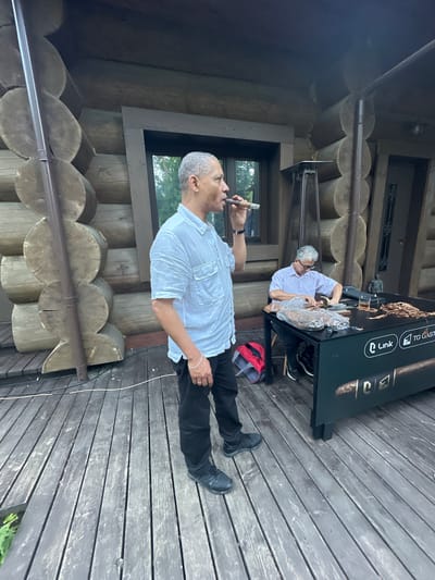 In Novoaleksandrovo, Russia, two men are on a wooden deck. One man, who is smoking a cigar, stands on the left side of the frame, wearing a white short-sleeved shirt and black pants. To the right, another man, wearing a light purple short-sleeved shirt and black pants, is seated at a black table. He appears to be working with tobacco leaves, and a cigar-making press is visible on the table. Dried tobacco leaves are spread across the table, along with a glass of amber liquid and a small statue. A red backpack rests on the ground near the seated man. The background features a rustic log cabin structure and a gas heater.