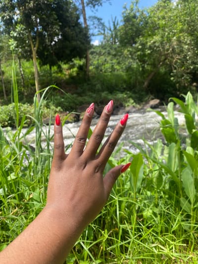 A close-up, dorsal view of a left human hand with dark brown skin is positioned in the foreground, extended slightly forward. The hand displays artificial fingernail extensions: the thumb, ring finger, and pinky finger are painted solid red, while the index and middle fingers feature a light pink base with distinct red wavy or swirl designs. The backdrop consists of a natural outdoor environment. Immediately behind the hand and in the midground, a flowing body of water, identifiable as a river or stream, displays visible white rapids and dark rocks. The foreground and banks of the water are densely covered with green vegetation, including tall grasses and broad-leafed plants. The background features additional green trees and lush foliage under a clear blue sky. The scene is situated in Kianyaga, Kenya.