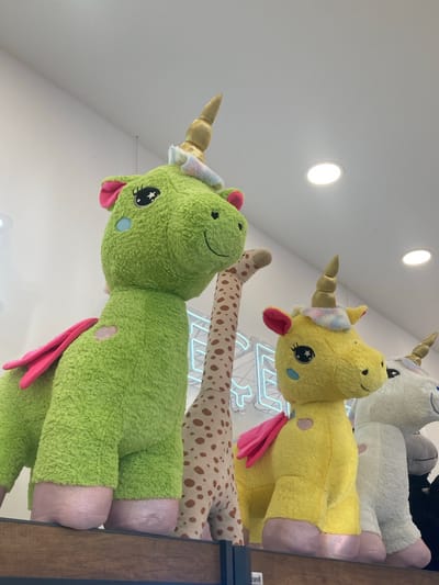 A low-angle, close-up shot shows a line of large, plush stuffed animals on a wooden shelf, illuminated by ceiling lights. In the foreground, a bright green unicorn with pink wings and hooves is positioned to the left. To its right, a spotted giraffe stands tall. Further to the right, a yellow unicorn with pink wings and hooves is visible, followed by a white unicorn with pink hooves. Behind the animals, a bright neon blue sign with partially visible letters is mounted on a white wall. The overall scene appears to be indoors, likely in a retail environment, possibly a toy store or gift shop. There are no people or actions depicted in the image. The location is not discernible from the image itself.