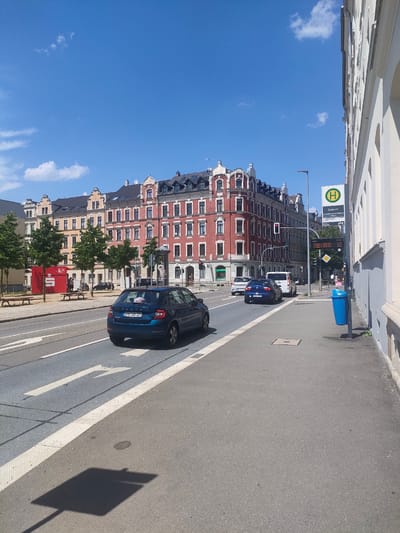 The image depicts a street scene in Chemnitz, Germany, on a sunny day with a clear blue sky. A multi-lane road runs horizontally across the frame, with vehicles moving in both directions. In the foreground, a blue Skoda hatchback is driving on the right side of the road, facing away from the camera. To the left of this car, a white van is visible. On the sidewalk to the right, a blue trash bin stands next to a bus stop sign with "H" for Haltestelle and "Zietenstr." as the stop name. A digital display board at the bus stop shows arrival times for various bus routes. Behind the road, there is a row of multi-story buildings with traditional European architecture, featuring red and yellow facades, pitched roofs, and numerous windows. Trees line the sidewalk in front of these buildings. A red structure with a white "S" logo is also visible on the left side of the frame, possibly a shop or service point. The scene appears to be a typical urban environment in Chemnitz.