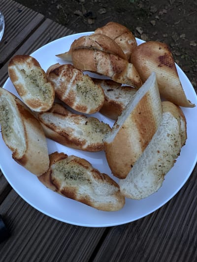 A white plate filled with toasted bread slices sits on a wooden surface, likely a table or deck. The bread appears to be baguette slices, some with visible herbs sprinkled on top and others with a more toasted, browned appearance. The slices vary in size and shape, suggesting they were cut from different parts of the baguette. The background shows a mix of dark soil and some dry leaves, indicating an outdoor setting. The lighting suggests it could be daytime, possibly overcast or in the shade, as there are no strong shadows. The wooden planks are weathered and gray, consistent with outdoor furniture. There is no visible text in the image. The overall scene depicts a simple food presentation, likely a side dish or appetizer.