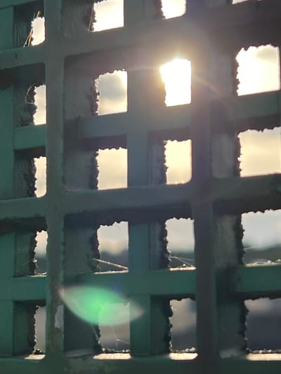 The image shows a close-up view of a green metal grate with square openings. Sunlight streams through some of the openings, creating bright highlights and lens flare. The background visible through the grate appears to be a sky with clouds, possibly during sunrise or sunset. The grate itself shows some wear and tear, with rough edges around the openings. The overall impression is one of light filtering through a barrier. The location is described as Terhillion, Ireland, though no specific landmarks or context to confirm this are visible in the image itself.